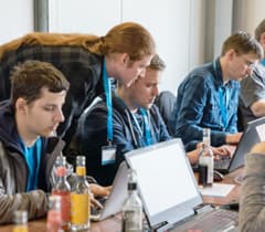 A group of developers is working together at a table, focused on their laptops. One person is leaning over to assist another developer.
